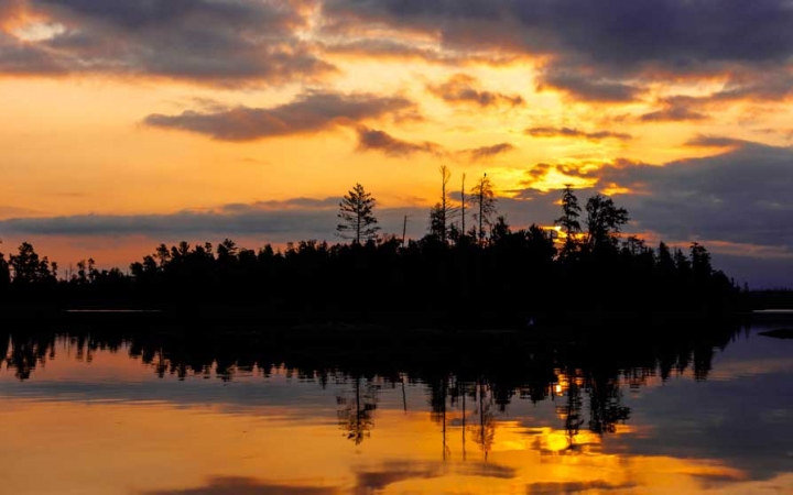 the sunset is reflected on a calm lake alongside a tree-lined shore